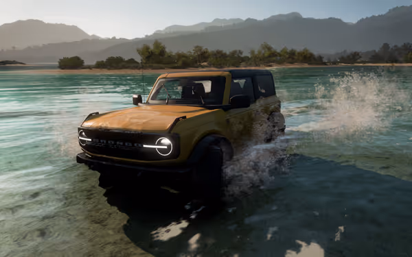 A yellow SUV drives through shallow water near trees, mountains, and a bright sky.