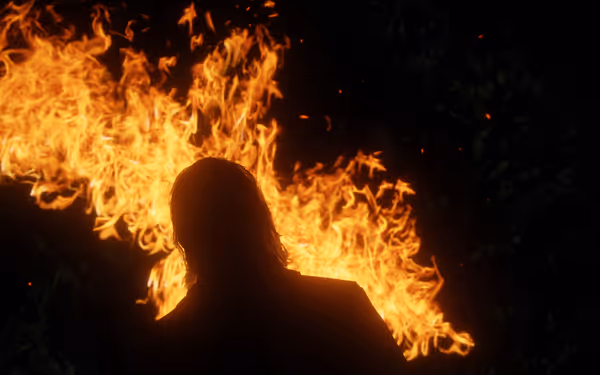 A male character is standing in front of flames at night in the forest.