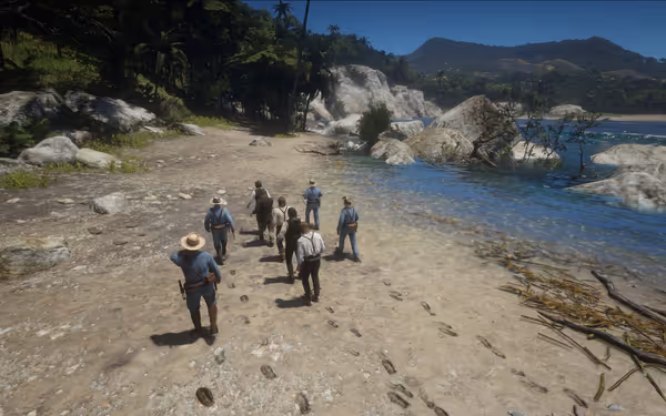 A group of people walks along a sandy beach near clear blue water, surrounded by lush greenery and rocky terrain.