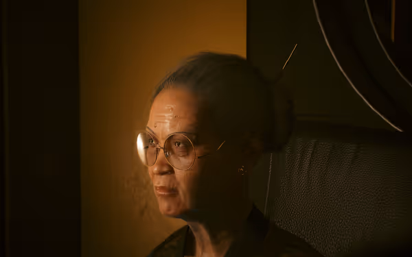 An elderly woman with glasses and pinned-back hair sits in a dimly lit room.
