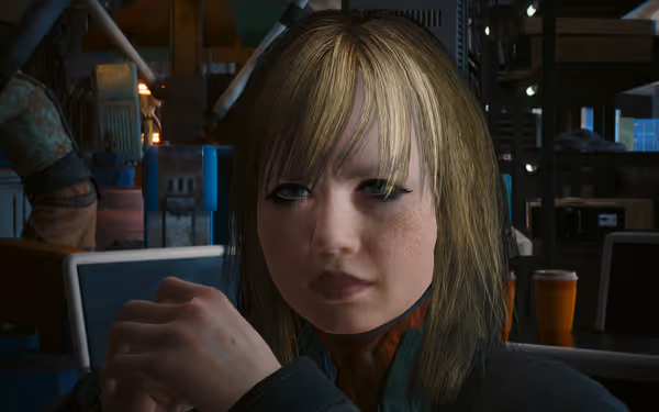 A blonde childlike woman with freckles and dark eyeliner sits indoors with hands clasped.