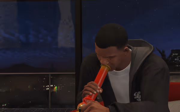 A man in a dark hoodie is smoking from a large red bong, overlooking a cityscape at night.