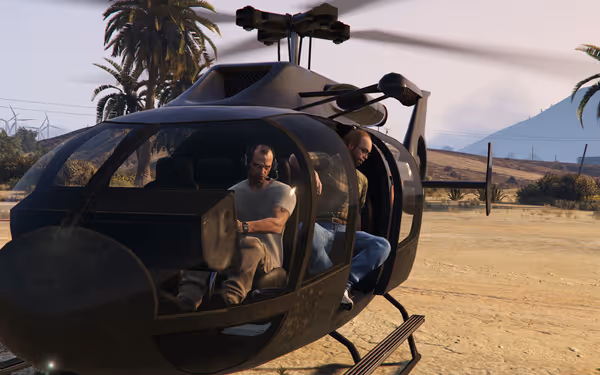 Two people sit inside a stationary black helicopter on a dirt field with palm trees.