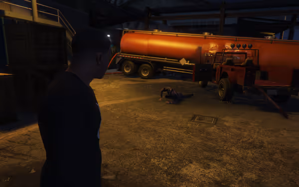 A man stands watching another man on the ground near a red truck and orange tanker.